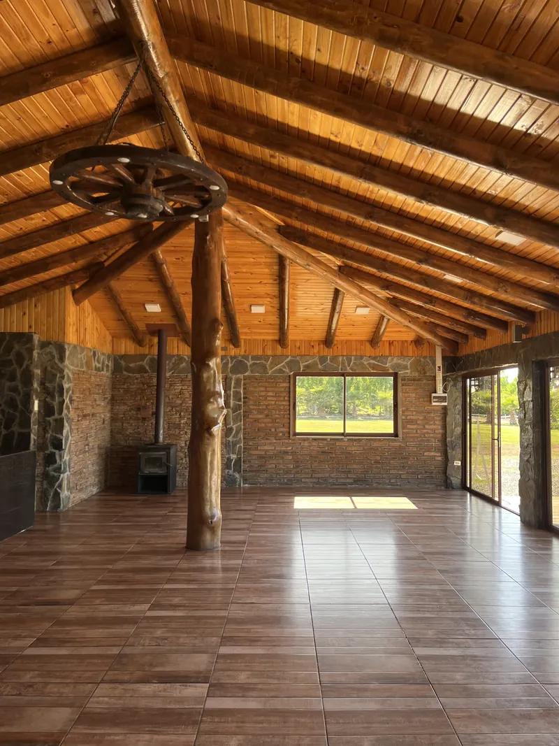 Interior amplio del quincho con techo de vigas de madera, muros de piedra y ventanales hacia el jardín