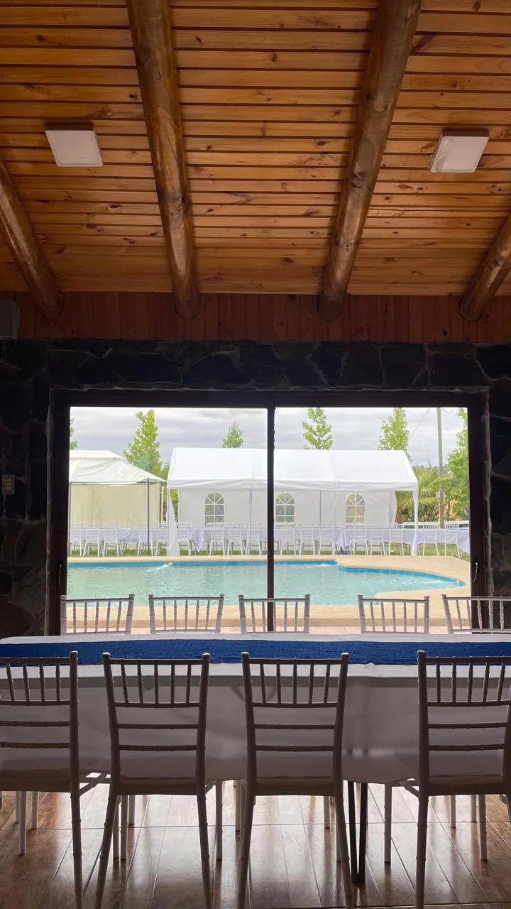 Vista desde el quincho: mesa vestida de blanco con sillas, y a través del ventanal la piscina y toldos con mesas preparadas